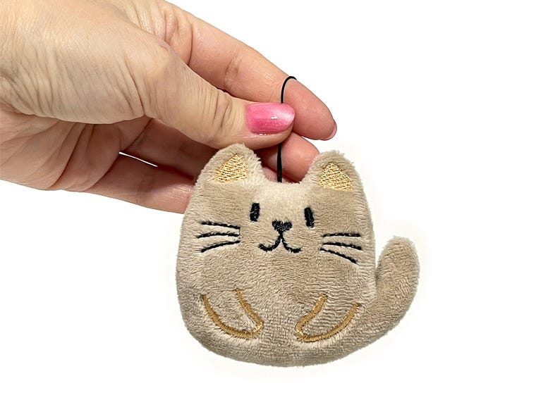 May include: A plush cat-shaped ornament in a light brown color. The cat has embroidered details, including a smiling face, whiskers, and paws. The ears are a darker shade of brown. A black string is attached for hanging.