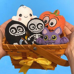Puede incluir: Una cesta de madera trenzada con una cinta amarilla que contiene cinco adornos de peluche: un fantasma blanco, una parca negra, un esqueleto blanco, una calabaza naranja y un murci&eacute;lago morado. Cada peluche tiene detalles bordados.