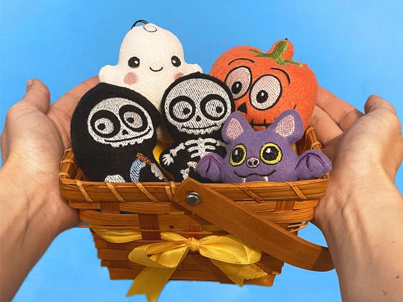 Puede incluir: Una cesta de madera trenzada con una cinta amarilla que contiene cinco adornos de peluche: un fantasma blanco, una parca negra, un esqueleto blanco, una calabaza naranja y un murci&eacute;lago morado. Cada peluche tiene detalles bordados.