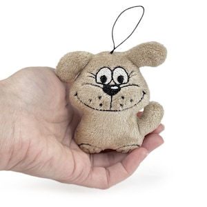 May include: A small, plush dog toy in tan with embroidered details. The dog has a smiling face, large eyes, and a black string loop for hanging. The toy is held in a hand against a white background.