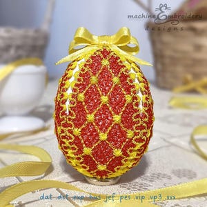 May include: A red and yellow embroidered Easter egg with a yellow bow. The egg is decorated with a diamond pattern and is sitting on a white surface.