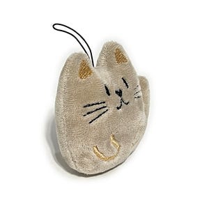 May include: A plush cat-shaped keychain in a light tan color. The cat has embroidered black whiskers and a smiling mouth, with gold accents on the ears and mouth. A black loop is attached for hanging.