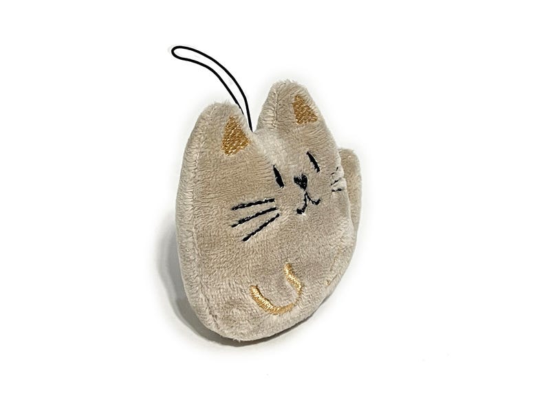 May include: A plush cat-shaped keychain in a light tan color. The cat has embroidered black whiskers and a smiling mouth, with gold accents on the ears and mouth. A black loop is attached for hanging.