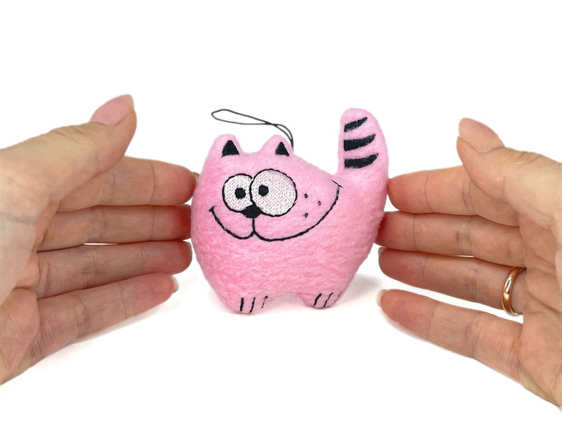 May include: A pink plush cat toy with black embroidered details. The cat has a smiling face, black stripes on its tail, and small black ears. A black string is attached for hanging.