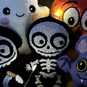 Puede incluir: Una colecci&oacute;n de cinco adornos de fieltro que incluye un fantasma blanco, una calabaza naranja, un esqueleto negro, un murci&eacute;lago morado y una figura negra con rasgos faciales blancos. Cada pieza tiene detalles bordados y peque&ntilde;as luces LED.