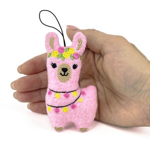 May include: A pink felt llama ornament with embroidered details. The llama has a floral crown and a garland of yellow and pink flowers. It has a black string for hanging.