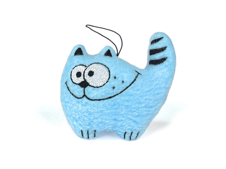 May include: A light blue cat-shaped plush toy with embroidered details. The cat has a smiling face, black whiskers, and a striped tail. It has a black string attached for hanging. The toy is made of soft material.