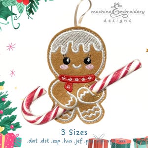 May include: A felt gingerbread man Christmas ornament with a red scarf and a candy cane. The ornament has white icing details and a smiling face. The text "machine embroidery designs" is at the top. The ornament is approximately 4 inches tall.