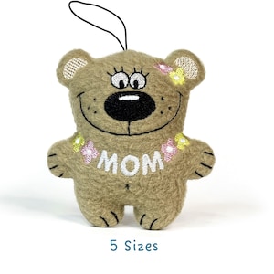 May include: A brown plush teddy bear with embroidered eyes, a black nose, and a smiling mouth. The bear is wearing a white necklace with pink and yellow flowers and the word "MOM" embroidered on the front.