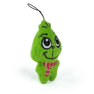 May include: Green plush ornament shaped like the Grinch with a red and white striped scarf. The ornament has embroidered eyes and a smiling mouth.