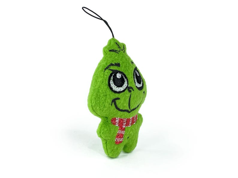May include: Green plush ornament shaped like the Grinch with a red and white striped scarf. The ornament has embroidered eyes and a smiling mouth.