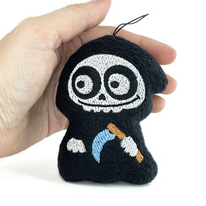Puede incluir: Un peque&ntilde;o adorno bordado negro, similar a un peluche, con forma de parca de dibujos animados. Presenta una cara de calavera blanca con ojos grandes, una boca sonriente y una guada&ntilde;a azul claro con mango marr&oacute;n sostenida por manos enguantadas.