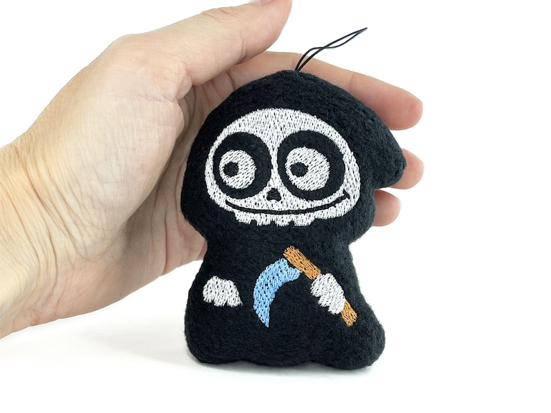 Puede incluir: Un peque&ntilde;o adorno bordado negro, similar a un peluche, con forma de parca de dibujos animados. Presenta una cara de calavera blanca con ojos grandes, una boca sonriente y una guada&ntilde;a azul claro con mango marr&oacute;n sostenida por manos enguantadas.