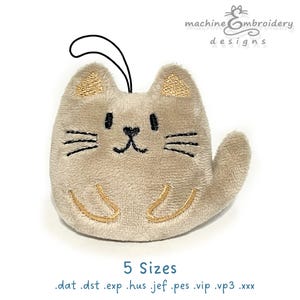 May include: A plush cat-shaped ornament in beige with embroidered details. The cat has black whiskers, eyes, and a nose, with gold accents on the ears and paws. A black loop is attached for hanging. The text "machine embroidery designs" and "5 Sizes" are also visible.