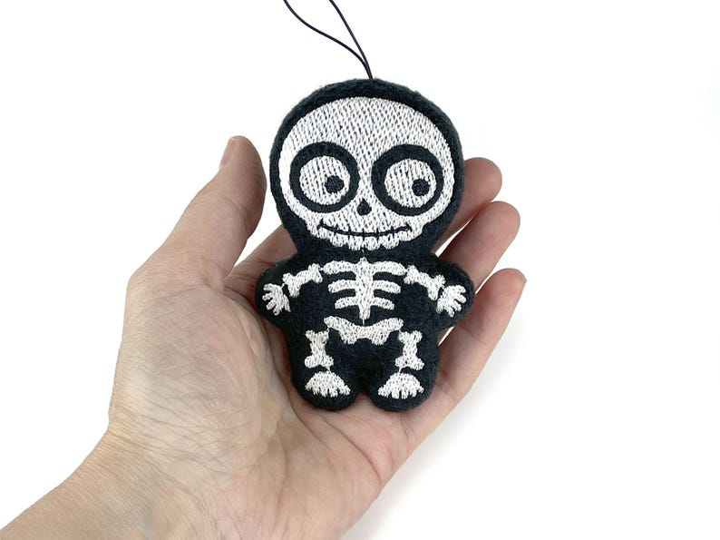 Puede incluir: Un peque&ntilde;o adorno de felpa negro con forma de esqueleto de dibujos animados. La figura presenta detalles bordados en blanco, incluyendo un cr&aacute;neo grande con ojos muy abiertos, una boca sonriente y una estructura &oacute;sea de costillas, brazos y piernas.