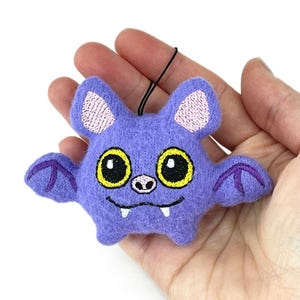 Puede incluir: Un adorno de murci&eacute;lago de fieltro morado con grandes ojos amarillos de pupilas negras, una peque&ntilde;a nariz rosa y dos colmillos blancos. El murci&eacute;lago tiene detalles de alas bordados en hilo morado oscuro, orejas rosa claro y un lazo negro para colgar.