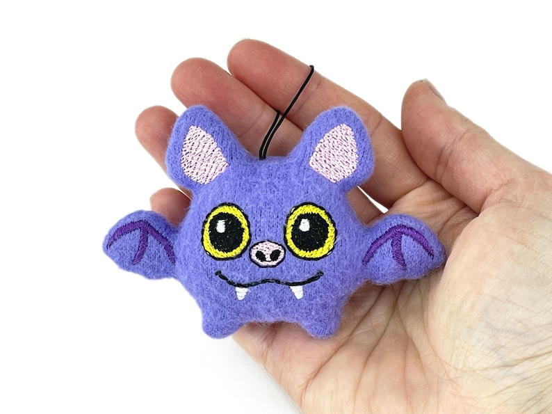 Puede incluir: Un adorno de murci&eacute;lago de fieltro morado con grandes ojos amarillos de pupilas negras, una peque&ntilde;a nariz rosa y dos colmillos blancos. El murci&eacute;lago tiene detalles de alas bordados en hilo morado oscuro, orejas rosa claro y un lazo negro para colgar.