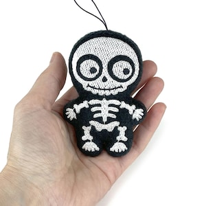 May include: A small plush skeleton toy with a black fabric body and white embroidered details. The skeleton has a smiling face with large eyes and a ribcage visible.