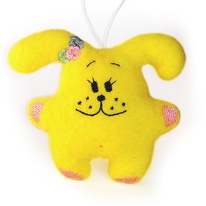 May include: A yellow felt toy dog with embroidered eyes, nose, and mouth. The dog has pink felt ears and paws and a flower crown on its head.