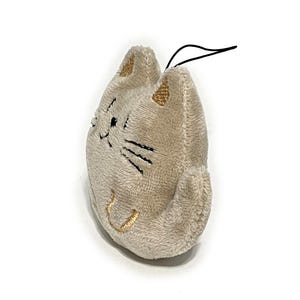 May include: A plush cat-shaped keychain in a light beige color. The cat has embroidered black whiskers and a smiling mouth. The ears are accented with gold thread. A black string is attached at the top.