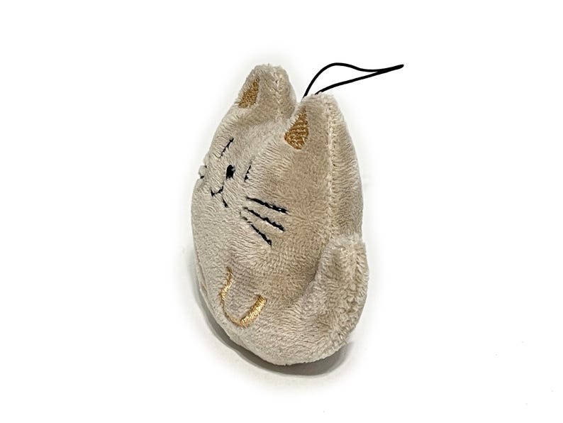 May include: A plush cat-shaped keychain in a light beige color. The cat has embroidered black whiskers and a smiling mouth. The ears are accented with gold thread. A black string is attached at the top.
