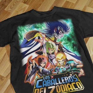 Puede incluir: Camiseta negra con un gráfico colorido de personajes de anime. El diseño incluye el texto "LOS CABALLEROS DEL ZODIACO" y presenta varias figuras en poses dinámicas sobre un fondo verde y azul.