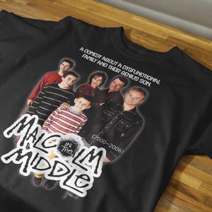 Puede incluir: Camiseta negra con un gráfico del elenco de la serie de televisión "Malcolm in the Middle". El diseño incluye el título del programa en una fuente estilizada, el elenco y los años 2000-2006.