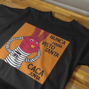 Puede incluir: Camiseta negra con un diseño de marioneta de conejo de dibujos animados. El conejo tiene una cabeza roja y brazos a rayas, sobre un cuadrado naranja. El texto en español dice "NUNCA HABÍA VISTO TANTA CACA JUNTA."