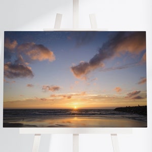 Może przedstawiać: Malowniczy zachód słońca nad plażą z molem w oddali. Niebo jest żywą mieszanką różowego, pomarańczowego i niebieskiego z puszystymi chmurami. Woda jest spokojna i odbija kolory nieba.