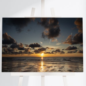 Może przedstawiać: Malowniczy zachód słońca nad plażą z ciemnymi chmurami na niebie i słońcem zachodzącym nad horyzontem. Woda jest spokojna i odbija pomarańczowe i żółte odcienie zachodu słońca.