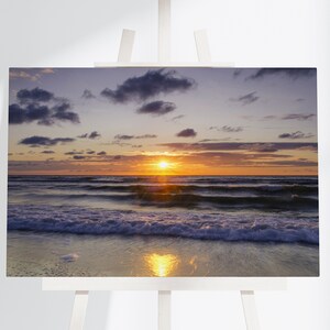 Może przedstawiać: Żywy zachód słońca nad spokojnym oceanem z falami delikatnie toczącymi się w kierunku brzegu. Niebo jest połączeniem fioletu, pomarańczy i różu z puszystymi chmurami.