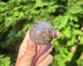 May include: A clear quartz sphere with inclusions of red, green, and black crystals. The sphere is approximately 5 cm in diameter.