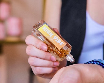 May include: A clear rectangular roller bottle labelled "HIGH VIBE" with a yellow and orange gradient. The bottle contains golden contents and is being applied to a wrist. A blue beaded bracelet is visible.