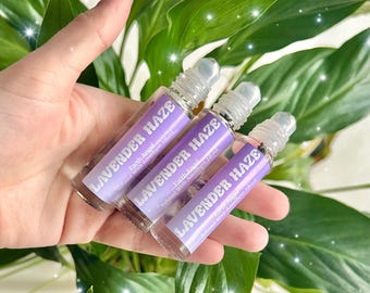 May include: Three clear glass roll-on bottles with purple labels. The labels read "LAVENDER HAZE" and "Earth Angel". The bottles contain a clear liquid and are held in front of green leaves.