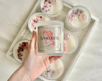 May include: A frosted glass candle with a white label that reads "Earth Angel Aphrodite Goddess of Love, Beauty, Passion Spell Candle 10 oz". The candle is surrounded by other candles in a white tray.