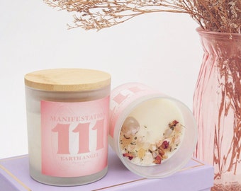 May include: Two pink glass candles with wooden lids. The candle on the left has the text "Manifestation 111 Earth Angel" on the label. The candle on the right has crystals and dried flowers embedded in the wax.