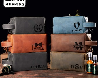 May include: A stack of personalized leather toiletry bags in various colors, including gray, brown, and blue. Each bag features custom text and designs, such as names and initials. The bags have a handle on one side and a zipper closure.