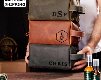 May include: Three leather toiletry bags in shades of brown and gray are stacked. The top bag is dark brown with the initials "DSP". The middle bag is tan with the initials "LM". The bottom bag is gray with the name "CHRIS". The bags have a handle and zipper.