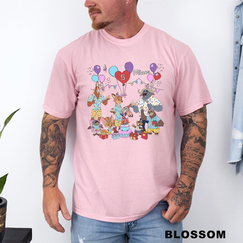 A man wearing a pink t-shirt with a colorful graphic design depicting a birthday party scene with balloons, presents, and various cartoon characters.