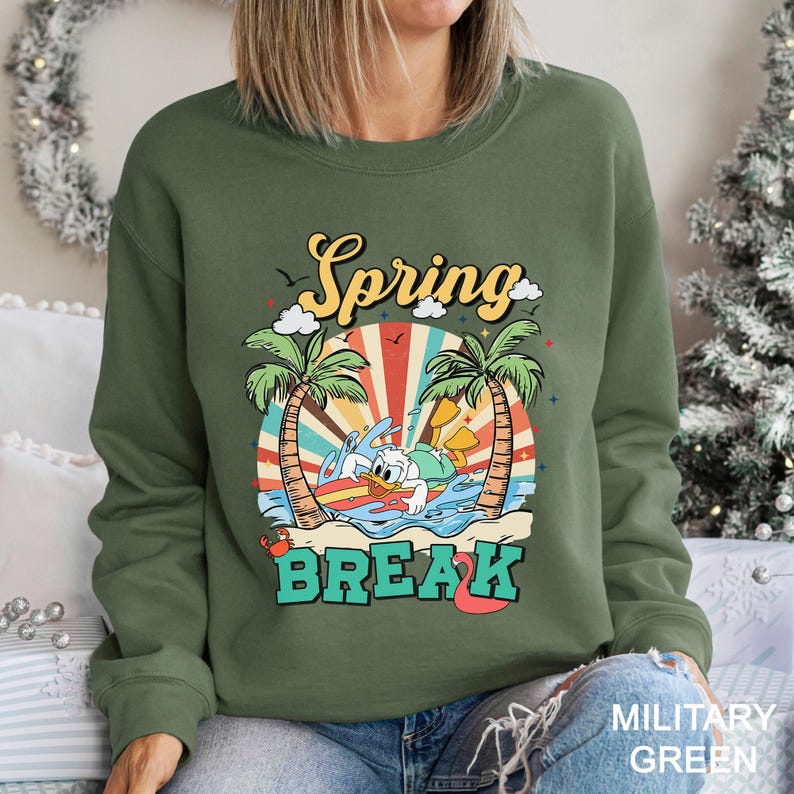 A woman wearing a green sweatshirt with a colorful graphic design depicting a cartoon character surfing on a wave, surrounded by palm trees and a sunburst pattern.