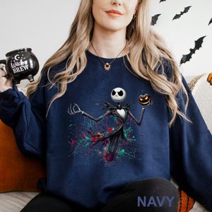 A woman wearing a navy blue sweatshirt with a graphic of a jack-o-lantern and a skeleton figure, holding a black mug with a "Witch Brew" design.