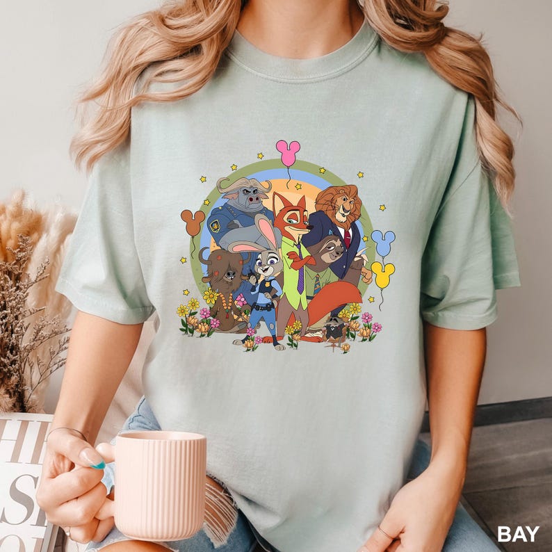 A woman wearing a t-shirt with a graphic of various cartoon characters, including a police officer, a fox, and other animals, along with flowers and balloons. She is holding a white mug in her hand.