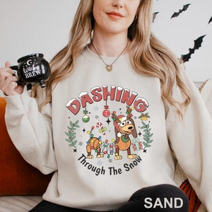A woman wearing a white sweatshirt with a graphic design of a cartoon dog and the text "DASHING Through The Snow" is holding a black mug with the text "Witch's Brew" on it.