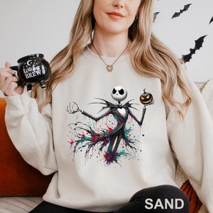 A woman wearing a white sweatshirt with a graphic of a jack-o-lantern and a mug with the text "Witch's Brew" printed on it.