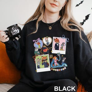 A woman wearing a black sweatshirt with a colorful graphic of cartoon characters on it, holding a black mug with the text "Witch Brew" printed on it.