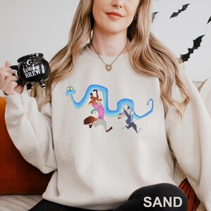 A woman wearing a white sweatshirt with a cartoon character design is holding a mug with a black and white design that says "Witch's Brew".