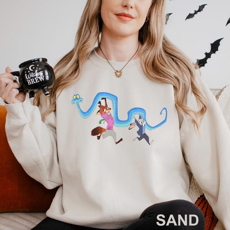 A woman wearing a white sweatshirt with a cartoon character design is holding a mug with a black and white design that says "Witch's Brew".