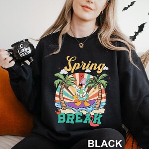 A woman wearing a black sweatshirt with a colorful graphic design and text, holding a black mug with a cartoon character on it.