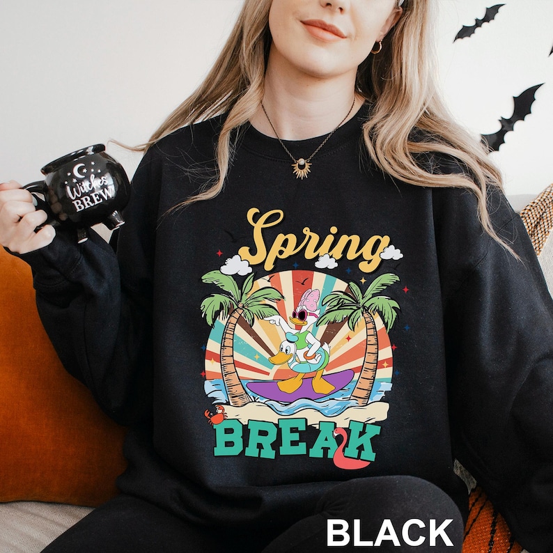 A woman wearing a black sweatshirt with a colorful graphic design and text, holding a black mug with a cartoon character on it.