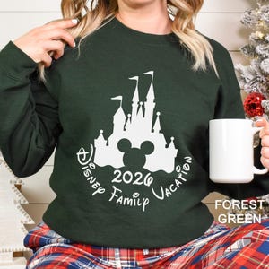A woman wearing a green sweater with a white castle and Mickey Mouse silhouette, holding a white mug, with a Christmas tree in the background.